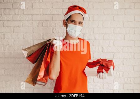 Junge fröhliche Lieferung Mann in medizinischen Gesichtsmaske, Schutzhandschuhe, santa Hut tragen Einkaufstaschen, Geschenkbox und Lächeln. Glücklicher Mann Kurier in Gesichtsmaske Stockfoto