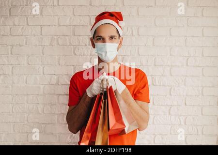 Happy man Kurier in santa hat, medizinische Gesichtsmaske, Schutzhandschuhe liefern Einkaufstaschen und Lächeln. Junge Lieferung Mann in Gesichtsmaske tragen Einkaufen b Stockfoto