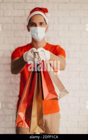 Glückliche Lieferung Mann in schützende Gesichtsmaske, medizinische Handschuhe, santa Hut tragen Einkaufstaschen. Junger Mann Kurier in Gesichtsmaske liefern Einkaufstaschen. Sicher Stockfoto