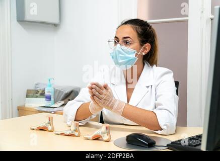 Eine junge Podologin mit einer Maske, die in ihrem Büro sitzt Als sie ihrem Patienten die Diagnose erklärt Stockfoto