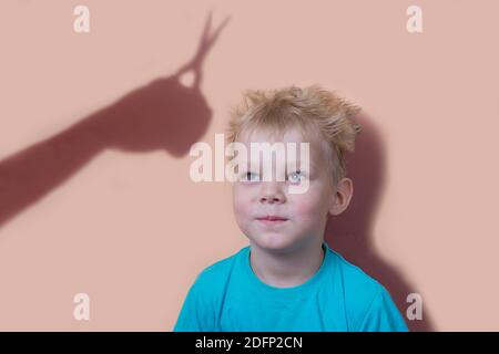 Der zottelige Junge schaut die Hand vom Bildschirm weg an.Haarschnitt. Der Schatten einer Hand mit Schere spiegelt sich an der Wand. Stockfoto