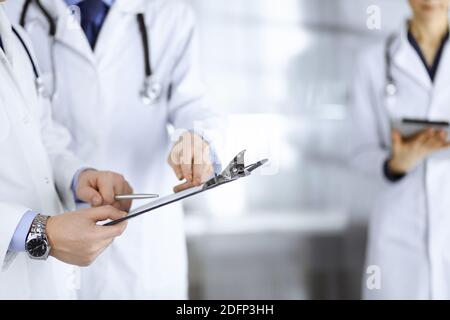 Unbekannte Ärzte, zwei Männer und eine Frau, diskutieren medizinische Untersuchungsresoults, während sie im Krankenhaus-Büro stehen. Ärzte, die Klemmbretter zum Auffüllen verwenden Stockfoto