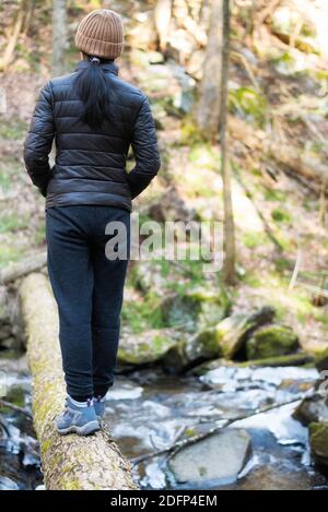 Eine Frau, die auf einem Baumstamm über einem fließenden, mit Eis und Schnee bedeckten Bach in der Dekanschlucht im Dorf connecticut von Falls am Wintertag steht. Stockfoto