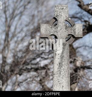 Ein schmutziges graues keltisches Steinkreuz in einem Friedhof. Das Kreuz ist schlicht ohne Markierung, und es ist etwas Moos darauf. Bäume sind im Hintergrund. Stockfoto