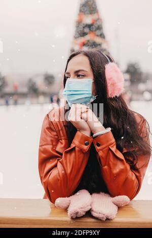 Schöne junge glückliche Frau in pelzigen Ohrenschützer, medizinische Gesichtsmaske haben Spaß auf Outdoor-Eisbahn. Fröhliche Mädchen in schützende Gesichtsmaske Skate auf Stockfoto