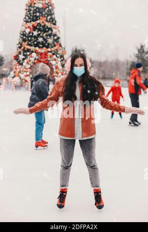 Attraktive fröhliche junge Frau in medizinischen Gesichtsmaske, rosa Ohrenschützer auf Weihnachten Eisbahn. Glücklich schöne stilvolle Mädchen in schützenden Gesicht Mas Stockfoto
