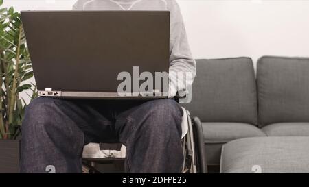 Junger Mann im Rollstuhl mit Laptop, der von zu Hause aus arbeitet, das Internet nutzt und auf den Bildschirm schaut. Stockfoto