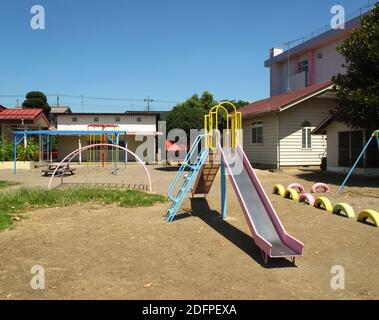 Ein traditioneller japanischer Grundschule Spielplatz mit Rutsche, Schaukel und Reifen mit Gebäuden im Hintergrund an einem sonnigen Tag im ländlichen Japan Stockfoto