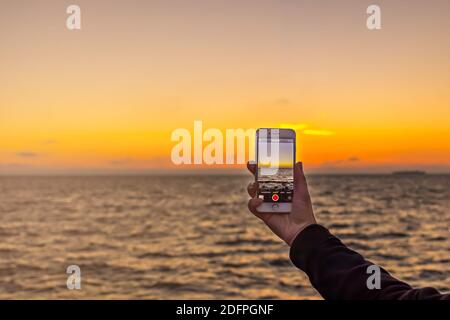 Die Hand einer Frau hält ein Smartphone und macht Fotos von einem Herbstuntergang. Stockfoto