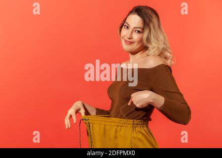 Portrait der schönen jungen erwachsenen Frau haben Willenskraft zeigt schlanke Taille in großen Trousen, erfolgreiche Gewichtsabnahme, Ernährung Konzeption. Innenaufnahme im Studio Stockfoto