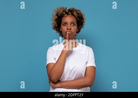 Nachdenkliche junge afroamerikanische Frau hält Kinn, sieht verwirrt beiseite, denkt über etwas nach. Studio auf blauer Wand. Stockfoto