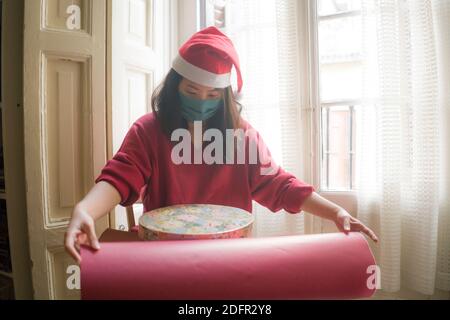 Frohe Weihnachten während covid19 - junge schöne und glückliche asiatische Chinesische Frau in Gesichtsmaske und Weihnachtsmann Hut Verpackung weihnachten präsent fröhlich Positi Stockfoto