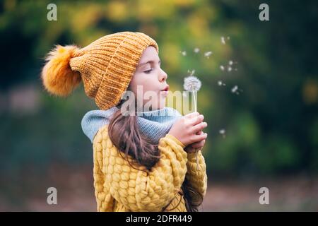 Kleines Mädchen, das im herbstlichen Park draußen spielt und Löwenzahn bläst. Herbstbäume im Stadtpark. Stockfoto