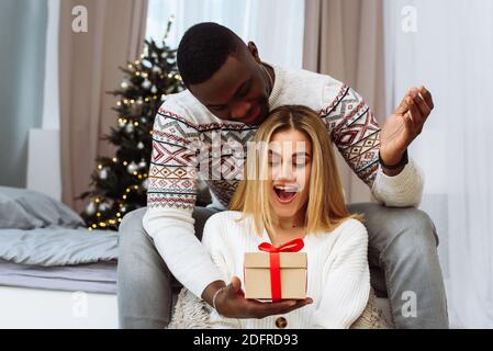 Eine junge kaukasische Frau erhält ein Geschenk von ihrem schwarzen Ehemann. Romantisches Paar Austausch Weihnachtsgeschenke Zu Hause. Stockfoto