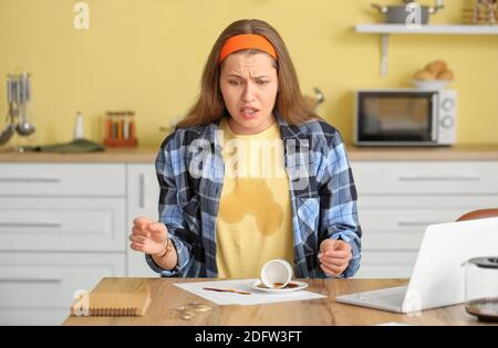 Gestresste junge Frau mit Kaffeeflecken auf ihrem T-Shirt und Verschütteter Kaffee in der Küche Stockfoto