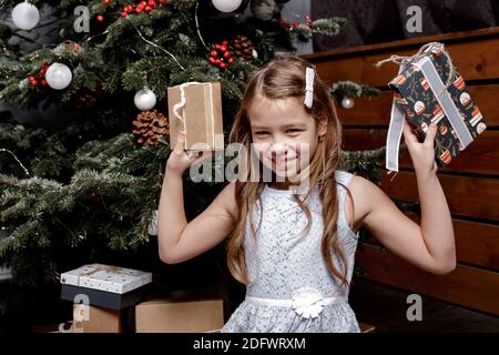 Glückliches Kind versucht zu erraten, was in ihren Geschenkboxen ist. Mädchen sitzt auf dem Boden in gemütlich dekorierten Wohnzimmer. Das Mädchen lacht und genießt das Geschenk. Stockfoto