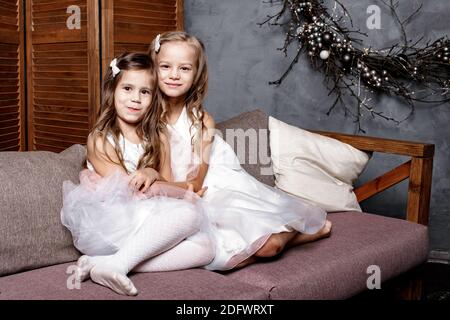 Zwei niedliche Schwestern in weißen Kleidern sitzen auf dem Sofa dicht beieinander und spielen zu Hause. Kleine Schwester umarmt ihre kleinste Schwester. Familie mit Chil Stockfoto