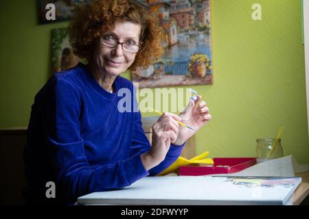 Glücklich reifen Künstler Frau Malerei auf Leinwand zu Hause Studio. Drücken Sie Ihre Kreativität aus. Positive ältere Frau hält Pinsel und Zeichnung auf Leinwand, während Stockfoto