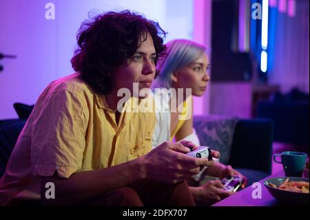 Glücklich Freund und Freundin spielen Videospiele und halten Joysticks zu Hause. Millennial Paar genießen Spiel, während auf dem Sofa sitzen und Ausgaben Stockfoto