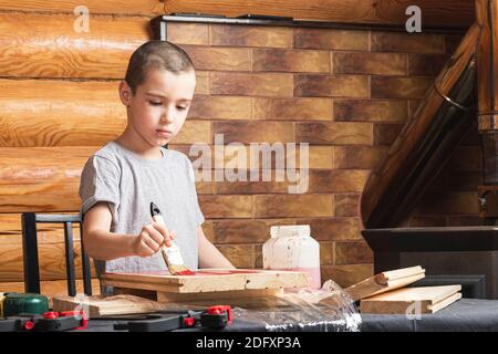 Ein Junge malt eine Holzfake mit einem Pinsel in rot in einem Landhaus. DIY Malerei der Wände. Junge, die zu Hause Renovierungen. Stockfoto