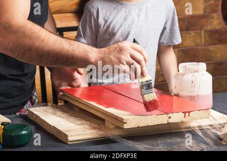 Vater und Sohn zusammen machen ein hölzernes Vogelhaus in der Werkstatt. Der fröhliche Vater mit dem kleinen Jungen bemalen das hölzerne Brett mit dem Pinsel in rot auf den Tisch Stockfoto