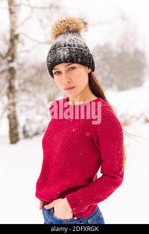 Ein junges schönes Mädchen in einem roten Pullover steht zwischen den Schneeverwehungen im Wald. Frau in rot in einem Park Winter, wenn es schneit. Stockfoto