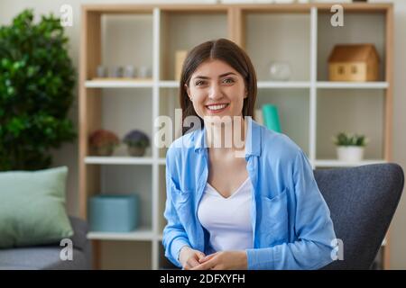 Lächelnde junge Frau in Freizeitkleidung sitzt auf Stuhl in Ihr Wohnzimmer mit Blick auf die Kamera Stockfoto