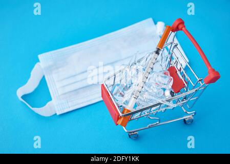 Spritzen und Ampullen mit Medizin im Warenkorb und medizinische Maske auf blauem Hintergrund. Kauf von Medikamenten Cocept Stockfoto