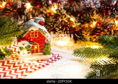 Weihnachtsspielzeug-ein Haus mit glühenden Fenstern und ein Schneemann in einer gemütlichen Umgebung. Neujahr, Serviette mit Zickzack, in den Lichtern der Girlanden. Urlaubsdekor Stockfoto