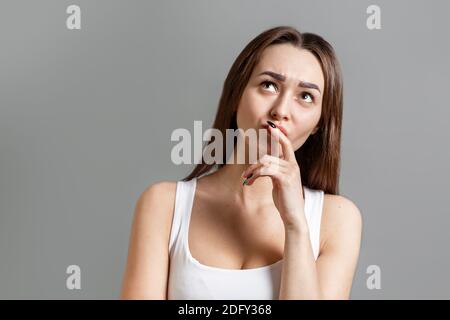 Konzept der Ideen- und Informationssuche. Porträt einer nachdenklichen jungen kaukasischen Frau, die mit einem Stirnrunzeln aufschaut. Grauer Hintergrund. Speicherplatz kopieren. Stockfoto