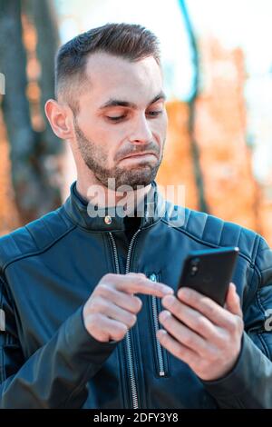 Jugendlich Kerl wurde überrascht mit schwarzen Smartphone im schönen Herbst Park. Schöner junger Mann mit Handy bei Sunny Day - mittlere Nahaufnahme Porträt Stockfoto
