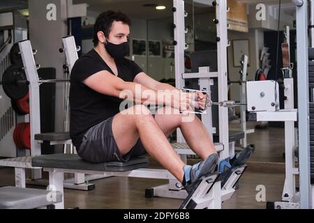 Fitness junger Mann trägt Schutzmaske mit Rudergerät im Fitnessstudio. Neue normale Übung. Stockfoto
