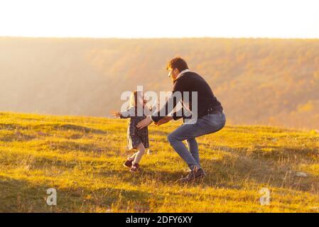 Familienurlaubung. Foto des Vaters, der seine Tochter, Wiese oder Park bei Sonnenuntergang fängt. Stockfoto