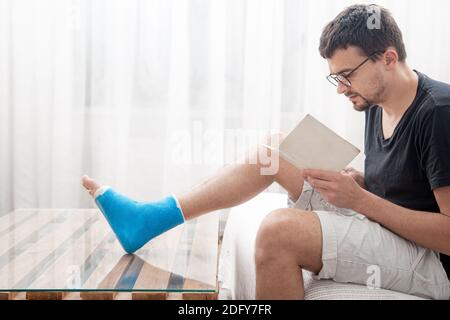 Ein junger Mann mit gebrochenem Bein in einer Besetzung liest Bücher auf einem hellen Hintergrund des Innenraums des Raumes. Stockfoto