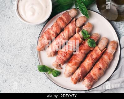 Ready-to-eat Schweine Würstchen eingewickelt in Speck an der Platte. Gebratene herzhafte Wurst in Speck umwickelt serviert frische grüne Blätter Basilikum mit Soße auf Hintergrund. Kopieren Sie Platz für Text. Top View oder flach. Stockfoto