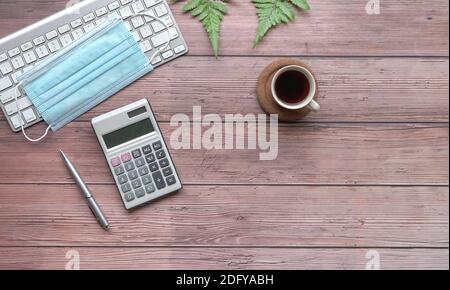 Holztisch mit Tastatur, Gesichtsmaske, Kaffee, Rechner und Grünpflanzen. Draufsicht. Stockfoto