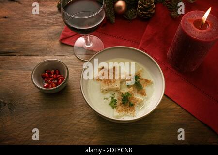 Weihnachtliche Sahnesuppe mit geröstetem Brot in Sternform, Petersiliengarnitur und Granatapfelkernen, Weinglas und brennender Kerze auf roter Serviette und dunkler R Stockfoto