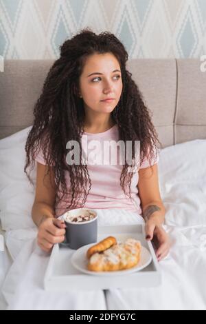 Frau frühstücken im Bett in hellen Hotel-Wohnung oder zu Hause. Fensterlicht Porträt junge Mädchen essen Croissant und Kaffee trinken. Stockfoto