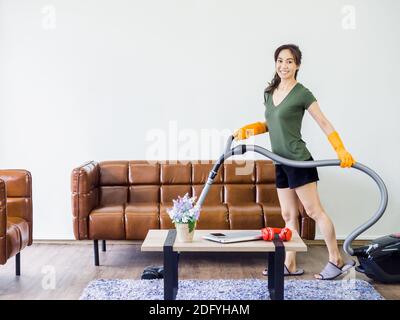 Junge asiatische Frau, Hausfrau in Freizeitkleidung und orange Gummihandschuhe mit Staubsauger, um den Boden im Wohnzimmer in der Nähe von braunem Ledersofa und zu reinigen Stockfoto