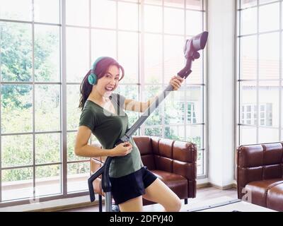 Junge schöne asiatische Frau, Hausfrau in casual Tuch Musik hören mit Kopfhörer und mit Staubsauger vorgeben Spaß mit Gitarre spielen im Leben Stockfoto
