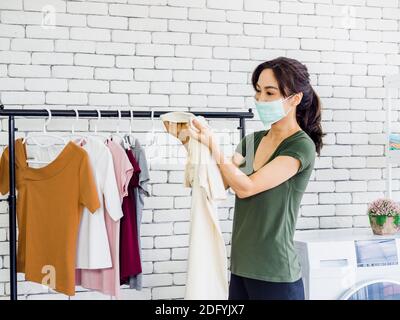 Junge schöne asiatische Frau, Hausfrau tragen Casual Tuch und Schutzmaske halten Hemd, Überprüfung schmutzigen Fleck auf sie nach dem Waschen vor dr Stockfoto