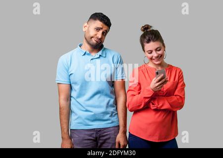 Portrait von lustigen umgekippt Mann in Freizeitkleidung mit Hände unten stehend, gelangweilt und genervt, wütend über Frau mit Smartphone, ihn zu ignorieren. isol Stockfoto