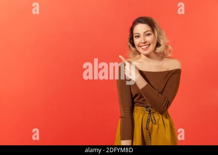 Porträt einer fröhlichen Frau mit blonden Haaren, die Finger beiseite zeigt, Kopieplatz für kommerziellen Text, leere Wand mit Idee Präsentation. Innen s Stockfoto