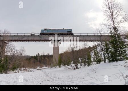 Eine Diesellokomotive fährt über eine hohe Betoneisenbahnbrücke über einen verschneiten Wald im Winter gegen einen bewölkten Himmel. Stockfoto