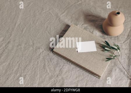 Hochzeit Schreibwaren Mock-up-Szene. Unbeschriftete Begrüßung, Visitenkarte und grüner Olivenzweig auf Buch. Keramikvase auf beigefarbenem Leinentischtuch Hintergrund Stockfoto