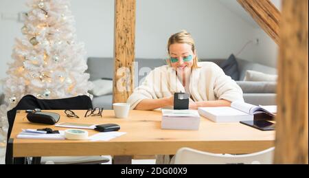 Junge Geschäftsfrau trägt kuschelig warmen Bademantel und kosmetische Augenflecken Arbeiten Sie in der Weihnachtszeit im Winter 2020 fernab von zu Hause corona-Virus Stockfoto