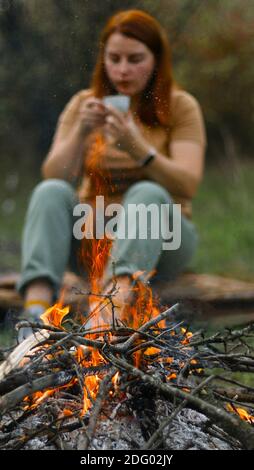 Lagerfeuer im Frühlingswald. Feuerkohlewald. Junges Mädchen trinkt ein heißes Getränk im Hintergrund, flache Tiefe des Feldes Stockfoto