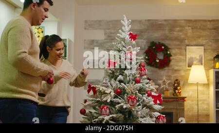 Paar mit passenden Kleidung schmücken ihre weihnachtsbaum für Winterferien. Dekorieren schönen Weihnachtsbaum mit Glaskugel Dekorationen. Frau und Mann in passenden Kleidern helfen verziertes Haus mit Girlande Lichter Stockfoto