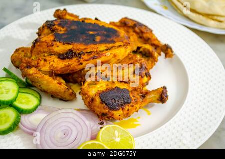 Schön platziert fertig verzehrfertige volle Huhn Tandoori auf einem weißen Teller zusammen mit geschnittenen Zwiebeln, Gurken und Zitrone Stockfoto