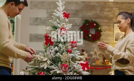 Paar mit passenden Kleidung schmücken ihre weihnachtsbaum für Winterferien. Dekorieren schönen Weihnachtsbaum mit Glaskugel Dekorationen. Frau und Mann in passenden Kleidern helfen verziertes Haus mit Girlande Lichter Stockfoto